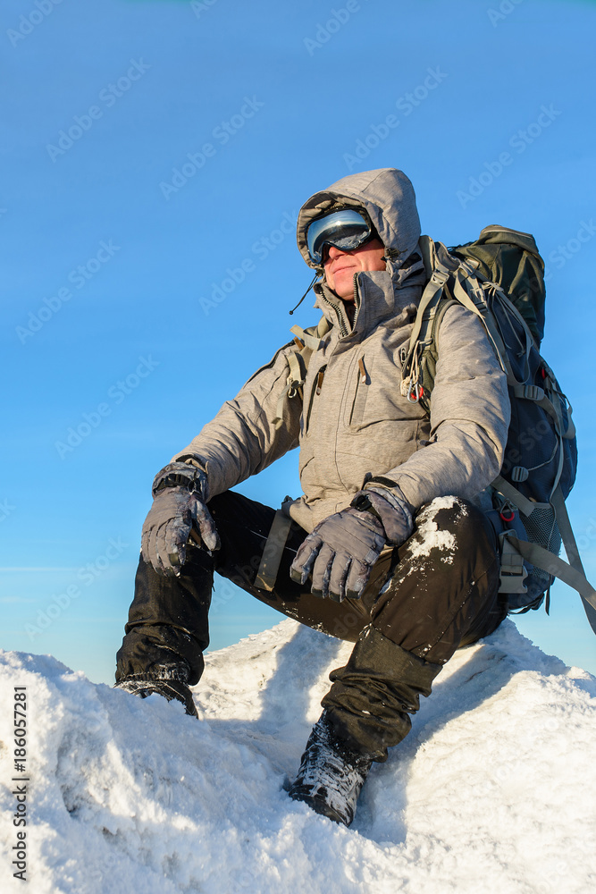 Hiker with backpack sitting and having rest on the top of a snow-covered rock over the winter mountains Hiker with backpack sitting and having rest on the top of a snow-covered rock over the winter mountains