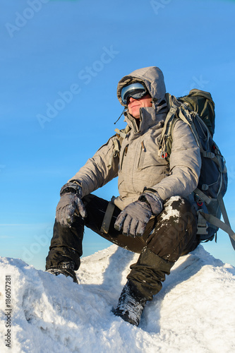 Hiker with backpack sitting and having rest on the top of a snow-covered rock over the winter mountains