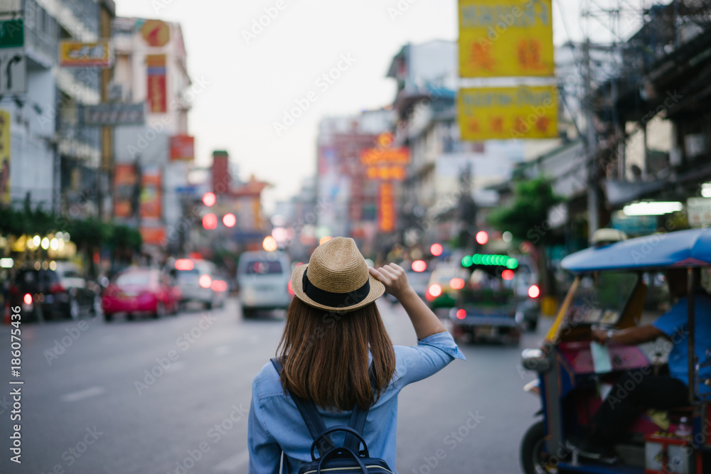 Young asian woman traveler with blue backpack and hipster hat looking night view on road with tuk tuk Thailand background at China Town Bangkok. Traveling in Bangkok Thailand