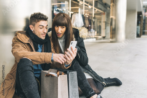 Young beautiful in love couple shopping around the street taking a break seating on the floor shopping on the mobile phone .