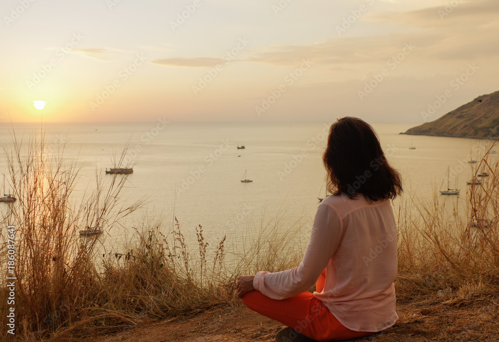 Young girl sitting in yoga lotus meditation position in front to seaside on the rocks an watching the sun goes down in a golden hour. sunset over sea.