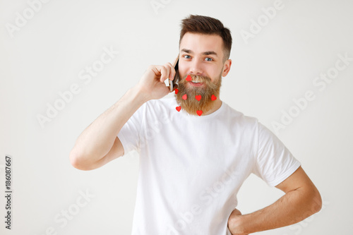 Positive pleasant looking bearded male model with long thick beard being satisfied to have phone conversation and relax after hard working day, enjoys spending time at home, isolated on white wall photo