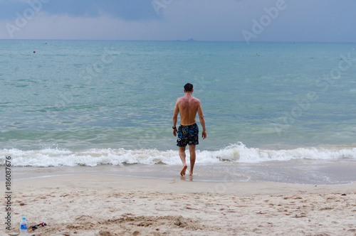 Man walking on the shore beach  sea in summer time. A charming man in shorts walking along the seashore. Waves and blue sea behind him. Lean physique and rightly muscular.