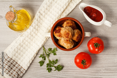 Fried meat cutlets in ceramic soup bowl, tomatoes, ceramic sauce boat with tomato sauce, branch of fresh parsley, kitchen towel and glass bottle with sunflower oil on grey wooden boards. Top view