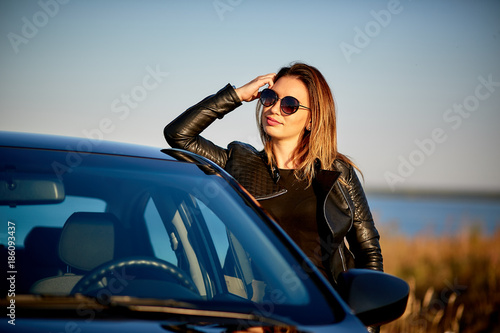 beautiful young woman in a leather jacket and sunglasses stands near the car