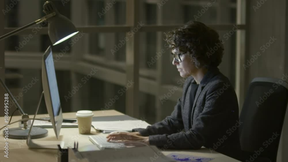 Side view of brunette businesswoman in eyeglasses typing on computer in the office at night Side view of brunette businesswoman in eyeglasses typing on computer in the office at night