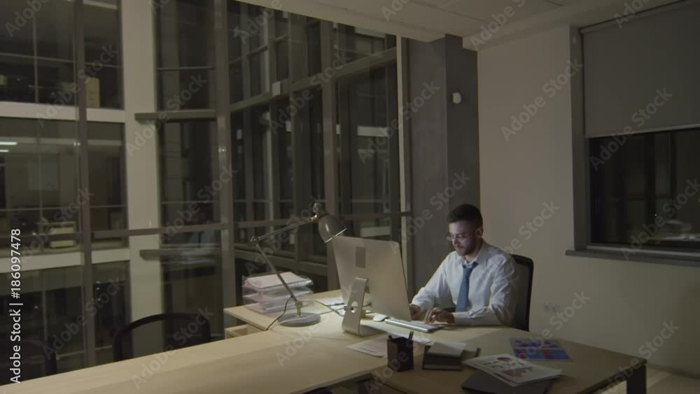 Young businessman sitting at desk in modern glass office and typing on computer in the night Young businessman sitting at desk in modern glass office and typing on computer in the night