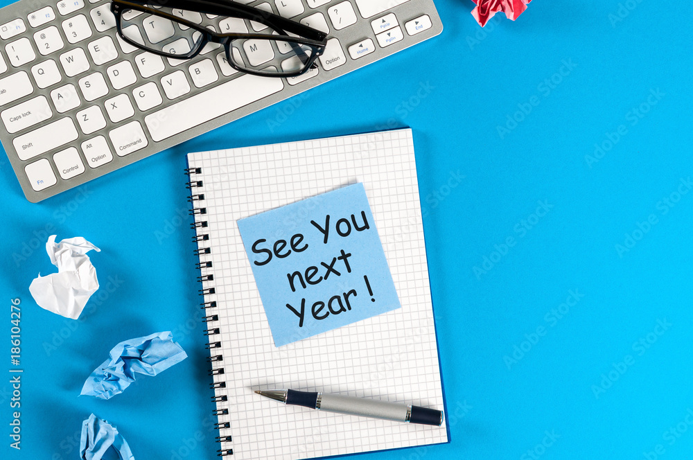 Business message See You Next Year written on notebook, with keyboard, office supplies and empty space at blue table in background