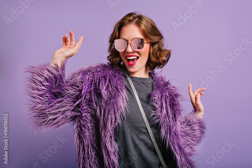 Stunning cute girl with curly brown hair posing with pleasure on purple background. Indoor photo of dancing young lady in trendy fur coat laughing to camera. photo