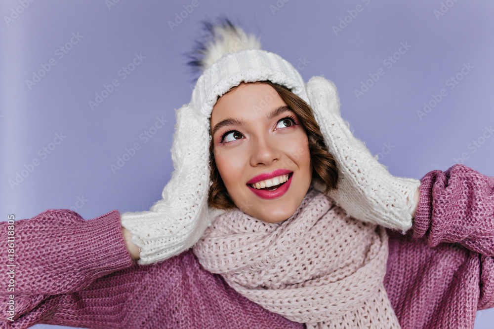 Close-up portrait of lovely girl with dreamy smile posing in white mittens. Fascinating young lady in knitted hat and scarf looking up and laughing on purple background.