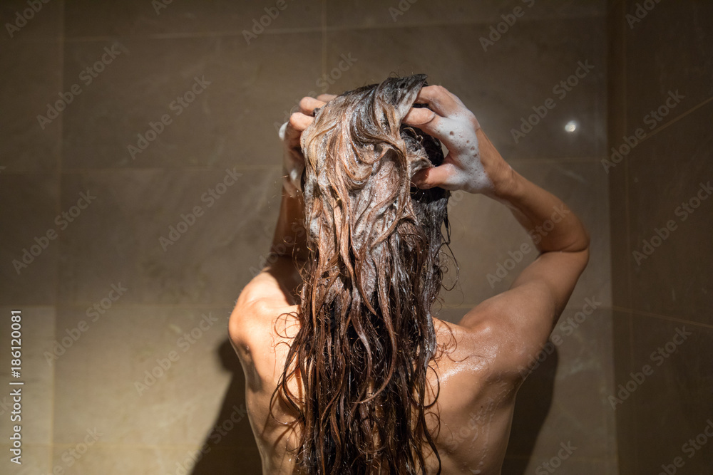 Naklejka premium adult woman from behind right washing her brunette long hair with shampoo and two hands in shower with brown marble tiles 