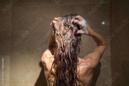 adult woman from behind right washing her brunette long hair with shampoo and two hands in shower with brown marble tiles
