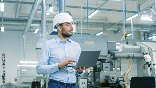 Tableau sur toile Chief Engineer in the Hard Hat Walks Through Light Modern Factory While Holding Laptop