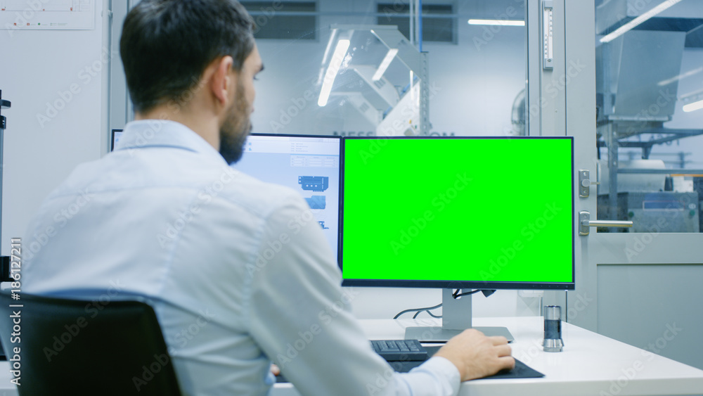 Engineer/ Technician Working on a Personal Computer with Two Displays, One Has a Green Screen Chroma Key Template Great for Mockup. Out of the Office Window Components Manufacturing Factory is Seen.