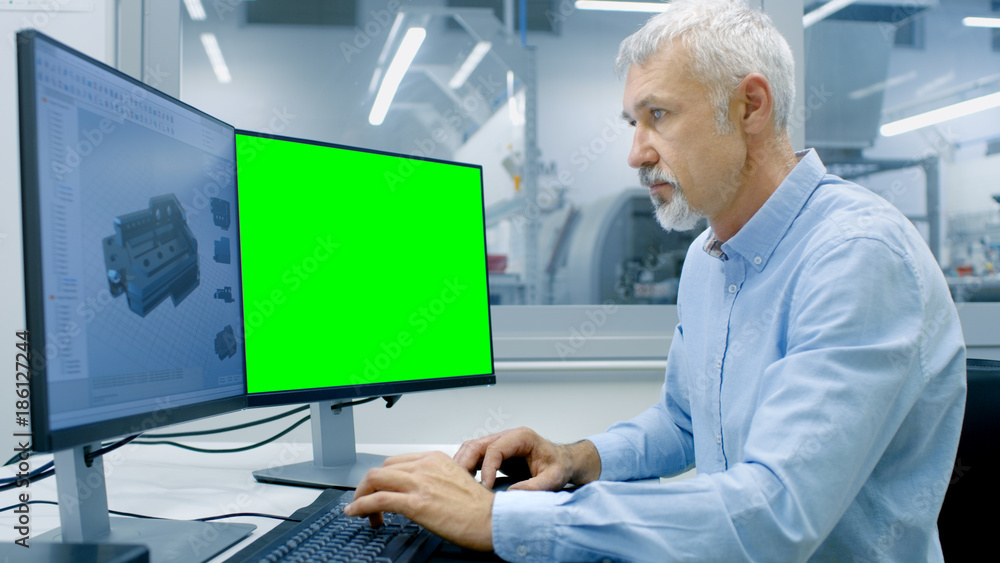 Engineer Working on a Computer with Two Displays, One has Chroma Key Great for Mockup Template and other Component in CAD Program. Out of the Office Window Components Manufacturing Factory is Visible. Engineer Working on a Computer with Two Displays, One has Chroma Key Great for Mockup Template and other Component in CAD Program. Out of the Office Window Components Manufacturing Factory is Visible.