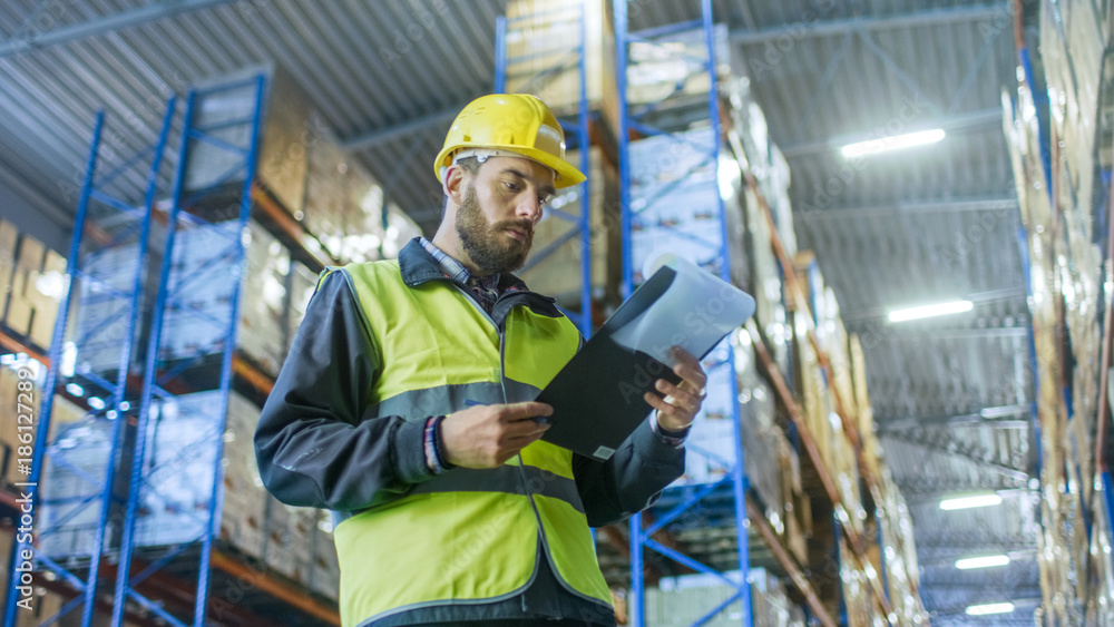 Overseer Wearing Hard Hat with Clipboard Fills in Forms in a Warehouse. He Walks Through Rows of Storage Racks with Merchandise. Overseer Wearing Hard Hat with Clipboard Fills in Forms in a Warehouse. He Walks Through Rows of Storage Racks with Merchandise.