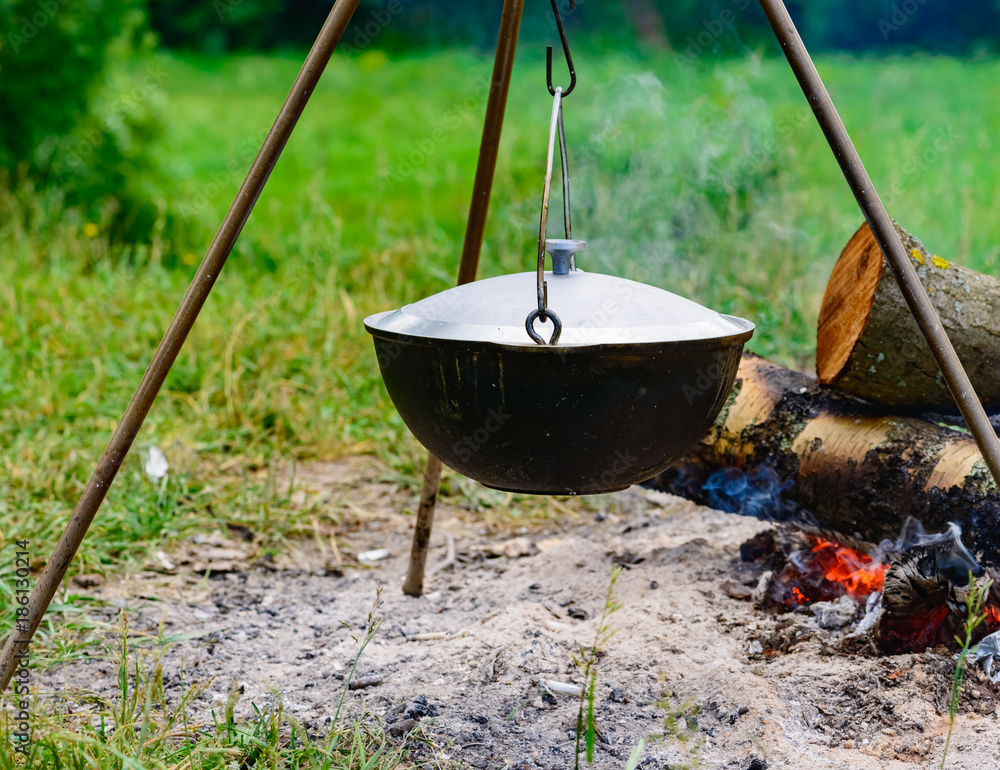 a camp-steaky sooty food bowl hanging on a tripod over a fire a camp-steaky sooty food bowl hanging on a tripod over a fire