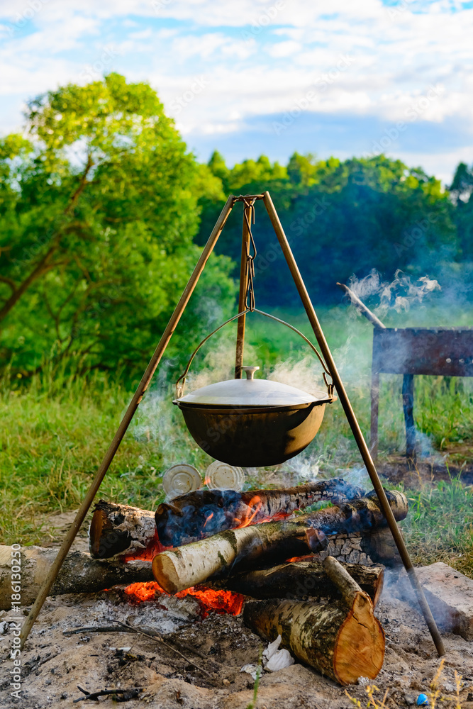 a camp-steaky sooty food bowl hanging on a tripod over a fire a camp-steaky sooty food bowl hanging on a tripod over a fire