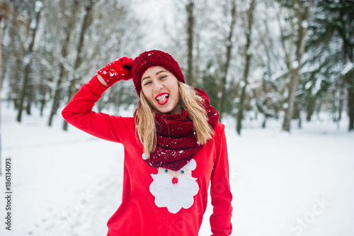 Blonde girl in red scarf, hat and santas sweater posing at park on winter day.