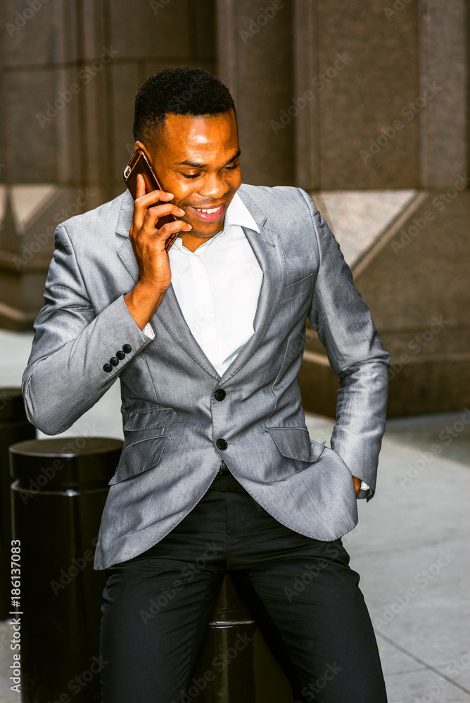 Happy African American Businessman working in New York. Wearing gray blazer white undershirt black pants young black man sitting on street outside office smiling talking on phone. City Daily Life foto...