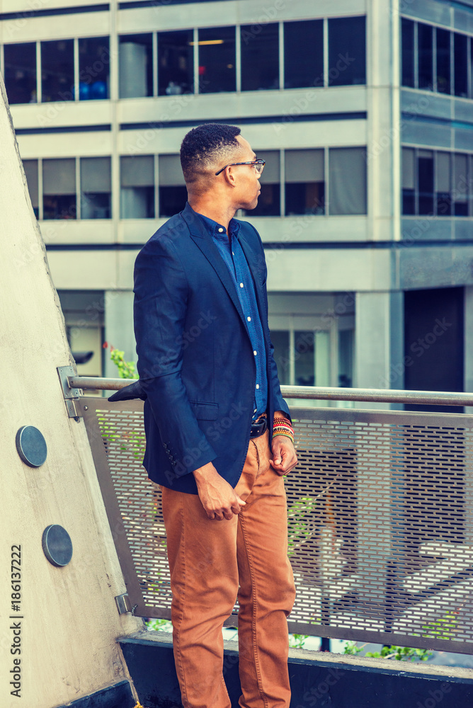 African American college student studying in New York, wearing blue jacket, brown  pants, glasses, bracelets, standing by railing on balcony, looking back,  facing street, thinking, lost in thought.. Stock Photo | Adobe