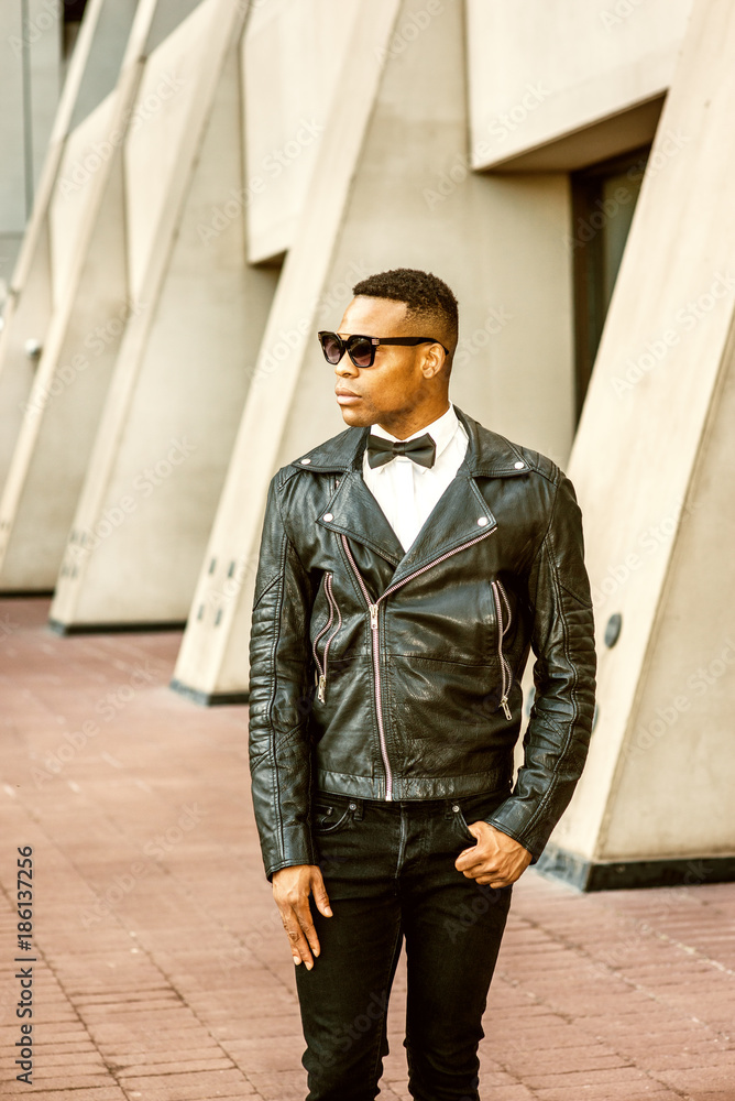 Man Urban Autumn/Spring Casual Fashion. Wearing black leather jacket, black  jeans, sunglasses, white undershirt, black bow tie, a young African American  guy standing on street in New York. City Boy.. Stock Photo