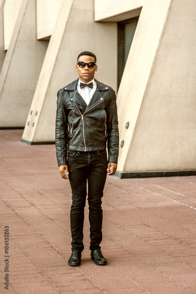 Man Urban Autumn/Spring Casual Fashion. Dressing in black leather jacket, jeans, leather shoes, white undershirt, black tie, wearing sunglasses, African American standing on in New York Stock