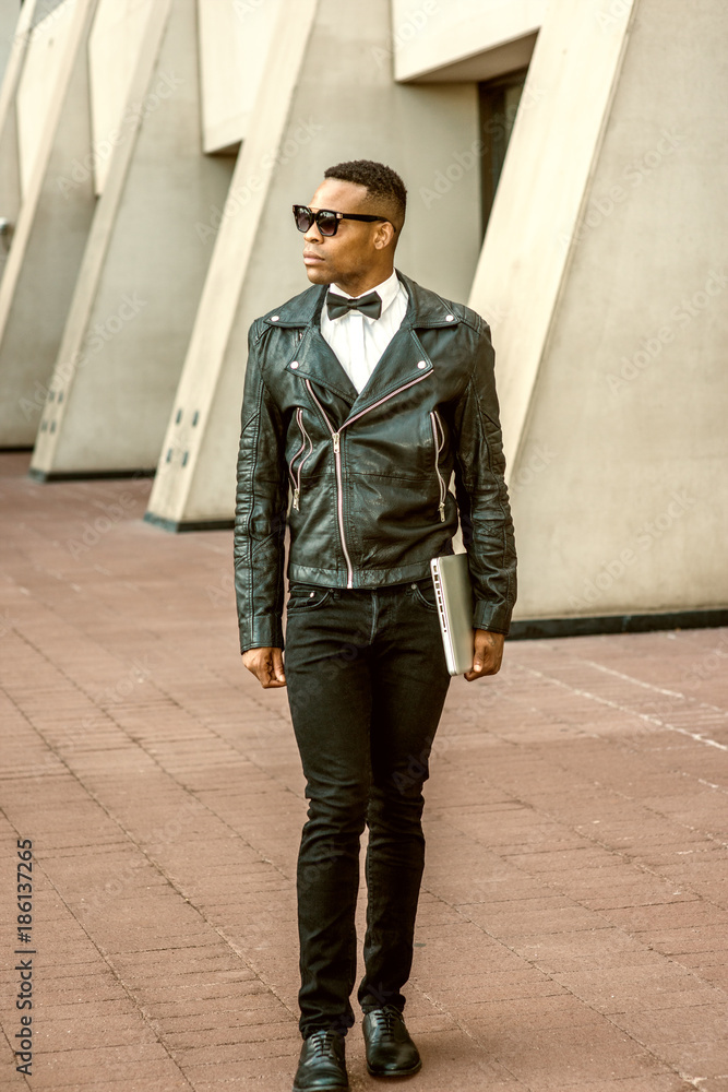 Man Urban Autumn/Spring Casual Fashion. Wearing black leather jacket, white  undershirt, black bow tie, jeans, sunglasses, carrying laptop computer,  African American guy walking on street in New York.. Stock Photo | Adobe
