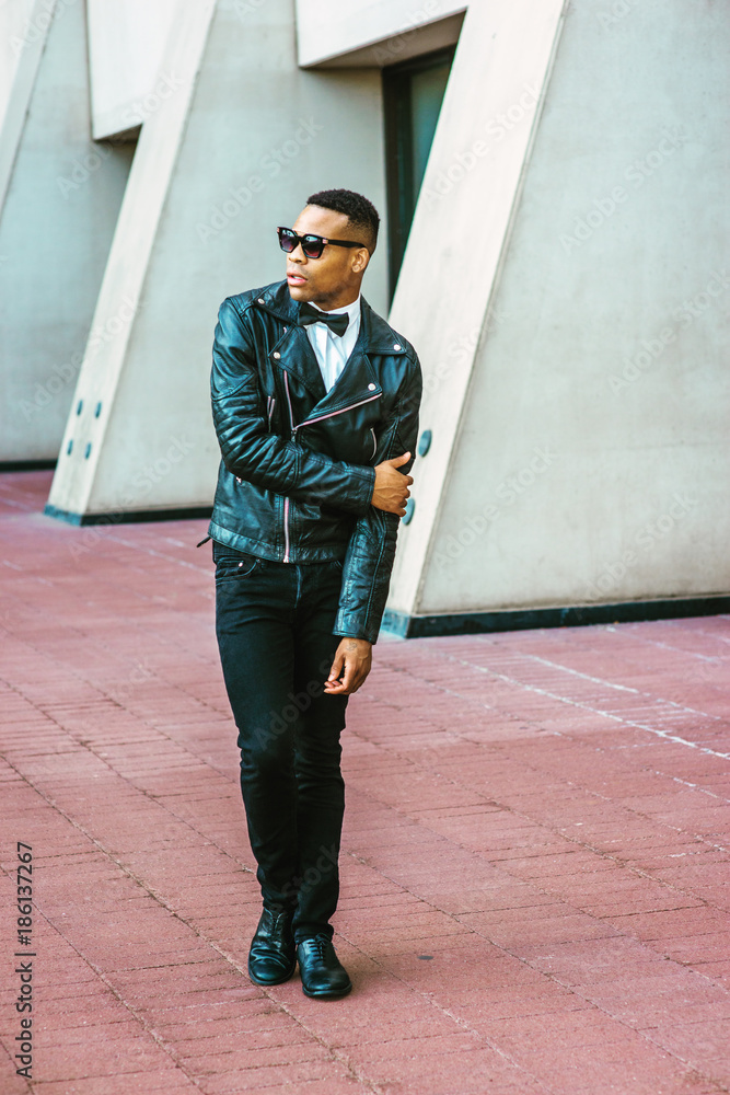 Man Urban Autumn/Spring Casual Fashion. Wearing black leather jacket, black  jeans, sunglasses, white undershirt, black bow tie, a young African American  guy standing on street in New York. City Boy. Stock Photo