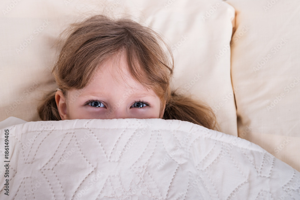 Little beautiful girl with blue eyes peeking out from under the blanket Little beautiful girl with blue eyes peeking out from under the blanket