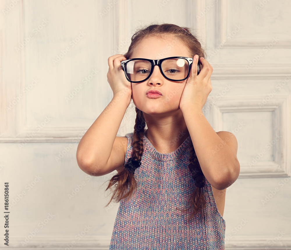Happy grimacing kid girl in fashion glasses with fun emotional face looking on blue studio background in colorful blouse. Closeup toned portrait Happy grimacing kid girl in fashion glasses with fun emotional face looking on blue studio background in colorful blouse. Closeup toned portrait