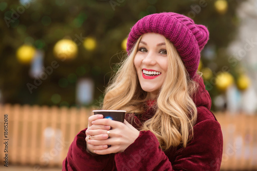 Happy young woman dressed in winter clothes and drinking coffee at the street