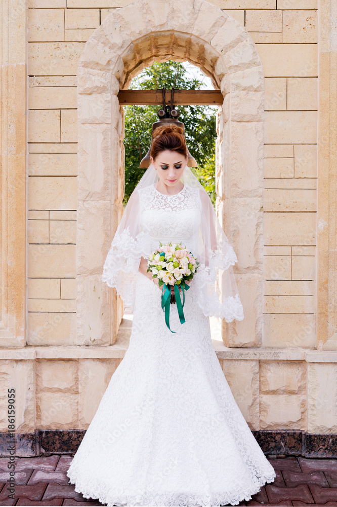 A beautiful bride in a wedding dress and a wedding bouquet in her hands stands outdoors against a stone arch with a bell. The concept of wedding happiness