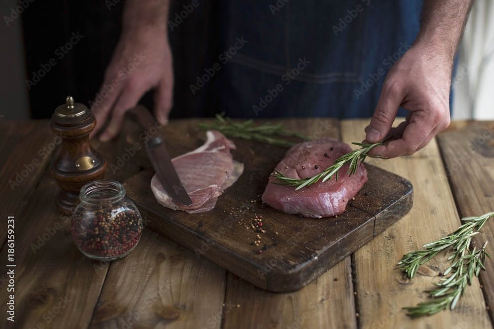 a man in a kitchen apron, meat on a board and a wooden table, rosemary, a knife and spices