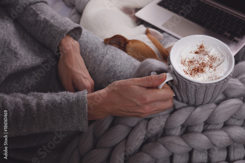 Woman in cozy home clothes relaxing at home, drinking cacao, using laptop. Comfy lifestyle.