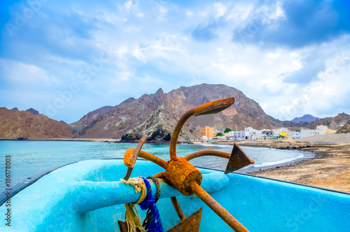 Old, rusty anchor resting on a fishing boat, blue sky and mountains.