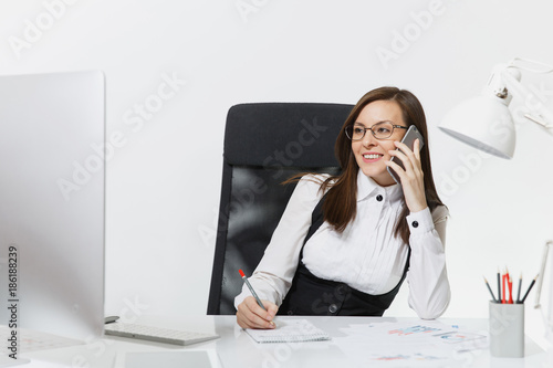Pretty smiling business woman in suit sitting at the desk, working at contemporary computer with document in light office, talking on mobile phone, conducting pleasant conversation on white background