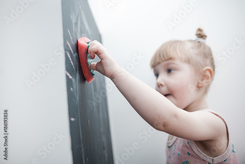 Little cute little girl paints with chalk on a blackboard at home, plays and wipes with a rag photo