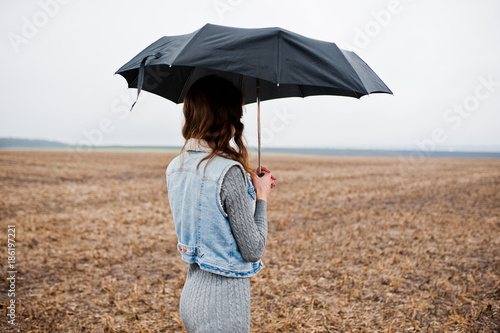 Portrait of brunette curly girl in jeans jacket with black umbrella at field.