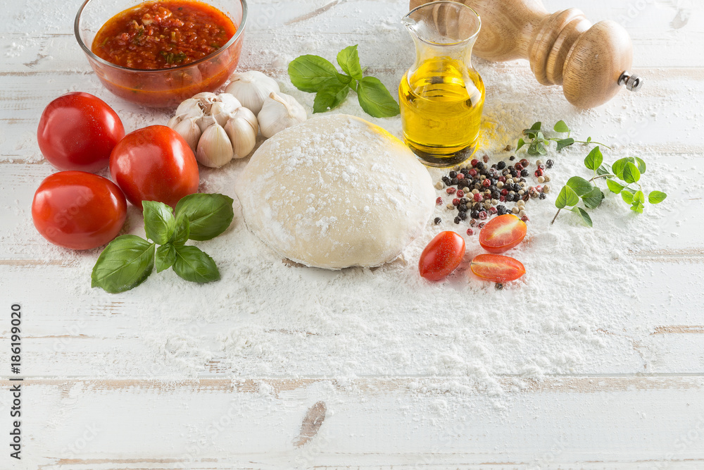 Fototapeta premium Food ingredients and spices for cooking, tomatoes, oil, pepper, salt, garlic, basil, oregano and delicious italian pizza dough with napkin, fork and spoon on white wood background. Top view.