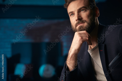 Portrait of pensive handsome man with dark hair  beard and mustache wearing dark jacket. Hand near face. Studio shot with blue lighted brick background with blurred clock and red barrel