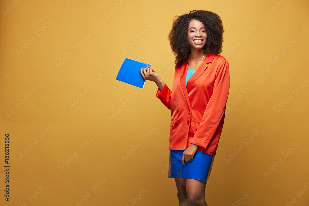 Portrait of young african girl with afro hairstyle. Smiling girl wearing orange jacket and violet latex skirt holds violet notebook in her hand and posing on yellow background. Studio shot. Portrait of young african girl with afro hairstyle. Smiling girl wearing orange jacket and violet latex skirt holds violet notebook in her hand and posing on yellow background. Studio shot.