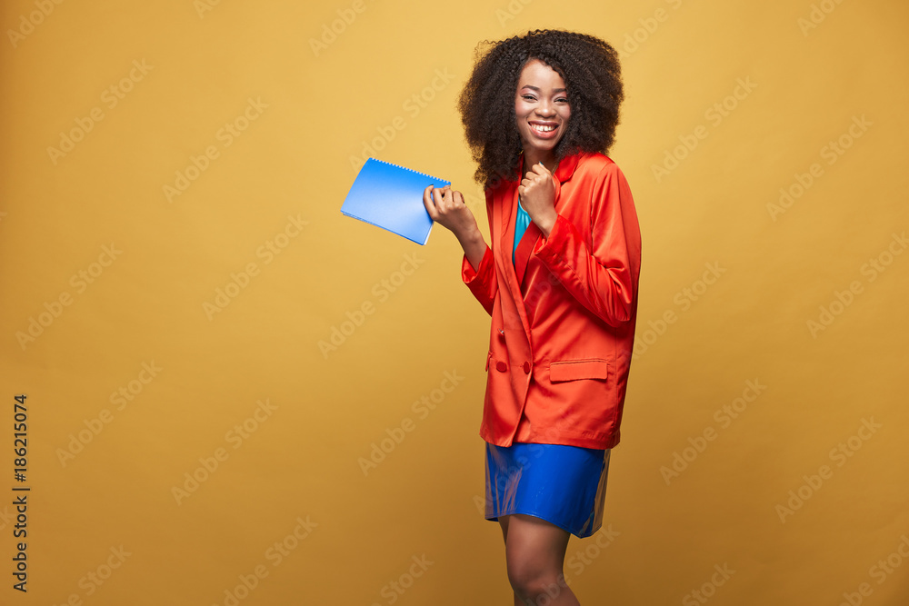 Bright portrait of young african girl with afro hairstyle. Smiling girl wearing red orange jacket and violet latex skirt holds violet notebook in her hand and posing on yellow background. Studio shot. Bright portrait of young african girl with afro hairstyle. Smiling girl wearing red orange jacket and violet latex skirt holds violet notebook in her hand and posing on yellow background. Studio shot.