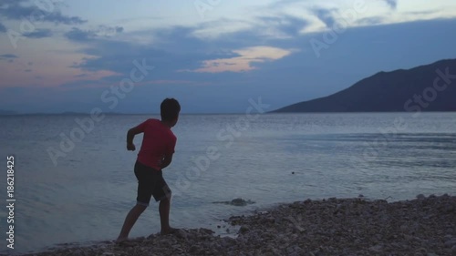 Slow Motion Of A Young Boy Playing, Skipping Stones By The Calm Blue Sea At Sunset.