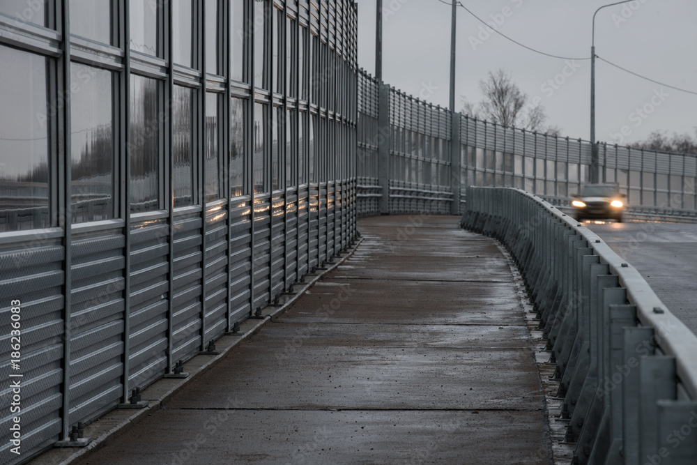 Protective noise barriers along the highway. A pedestrian road along the route. The car with the headlights on, shown in the blur Protective noise barriers along the highway. A pedestrian road along the route. The car with the headlights on, shown in the blur