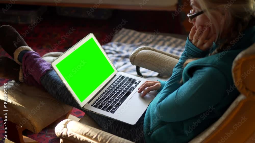 Beautiful backlit woman working on computer with green screen in a lazy boy chair with a dog curled up at her feet.