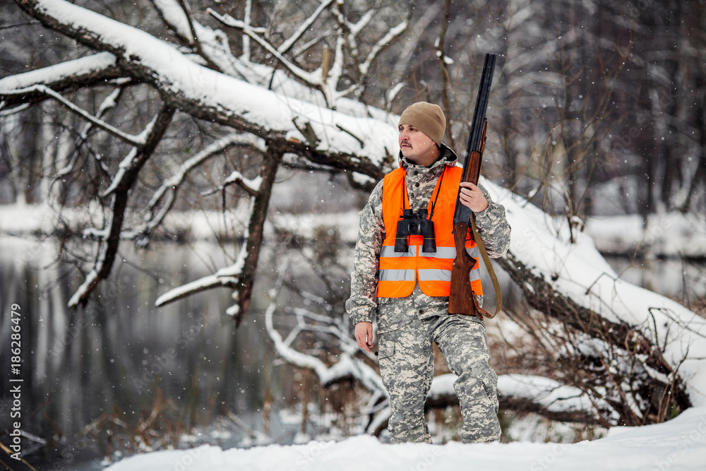 Male hunter in camouflage, armed with a rifle, standing in a snowy winter forest with duck prey