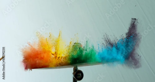 Colorful rainbow holi powder bounces off white canvas background in shockwave pattern of green blue pink and orange, slow motion, closeup