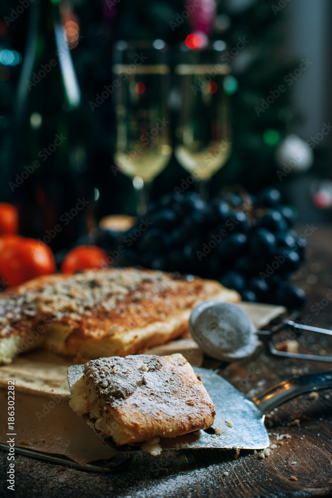 a festive New Year's table, cheese cake with chocolate, tangerines, grapes, biscuits, champagne and two glasses against the backdrop of a decorated Christmas tree with lights