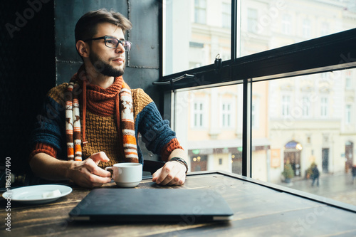 Vertical portrait of friendly bearded male freelancer in glasses dressed in sweater having a break with coffee, dreaming while sitting in loft studio near big window.